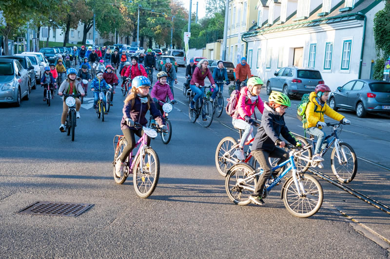 deti prvého stupňa základnej školy idú na bicykli vo veľkej skupine po chodníku do školy